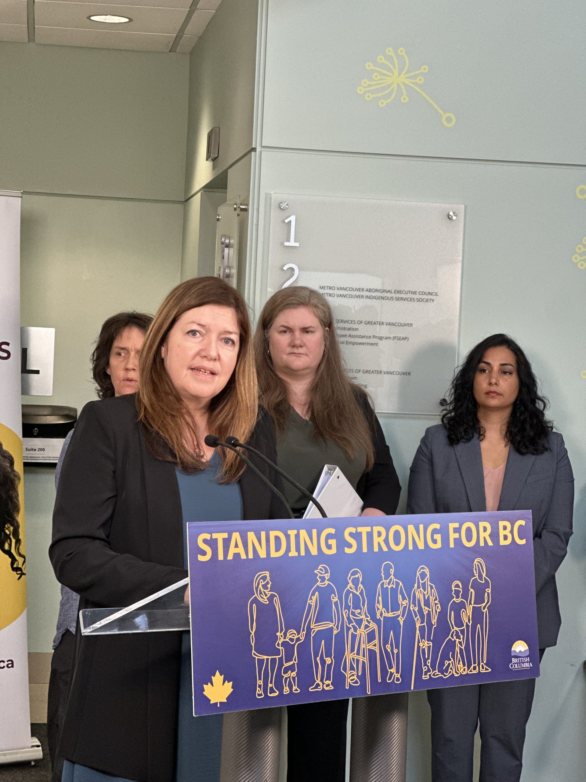Kim Stanton is standing at a podium delivering a speech. The podium features a sign for 'Standing Strong for BC', with the British Columbia logo.
